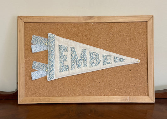 Fabric letter banner spelling 'EMBER' in a wooden frame on a cork board.